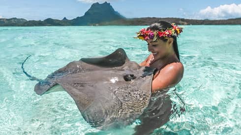 Woman wearing pink, red and yellow floral crown stands in the ocean holding a large manta ray in her arms