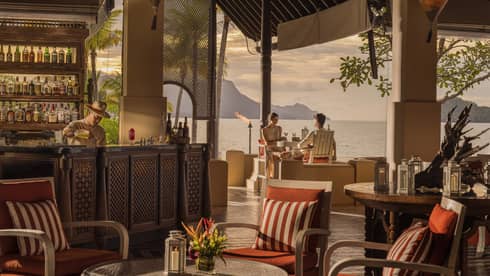 Two guests sips cocktails at open-air bar with ocean views while bartender makes drink on nearby bartop