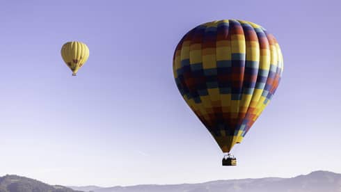 Close-up of a multi-coloured hot-air balloon close to the ground; a second yellow and red balloon floats higher above it.