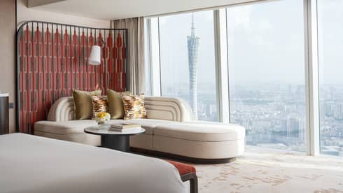 Hotel room with ivory-coloured corner sofa next to wall of windows, overlooking cityscape