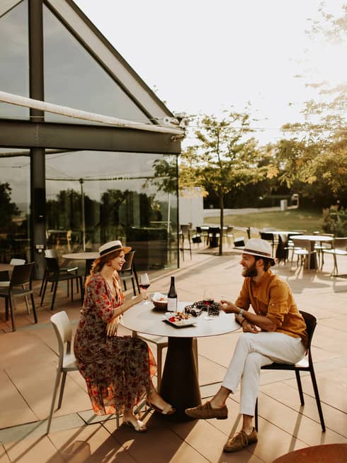Couple wearing hats sit at outdoor table with plate of food, drinking red wine