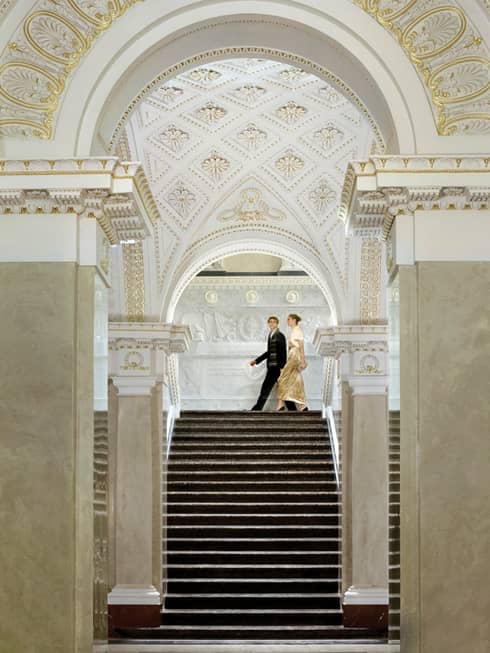 Woman in gold evening gown with fur jacket and man in suit walk at top of marble staircase