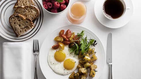 Breakfast plate with sunny-side-up eggs, cherry tomatoes, greens, and roasted potatoes, alongside toast, raspberries, grapefruit juice and coffee.