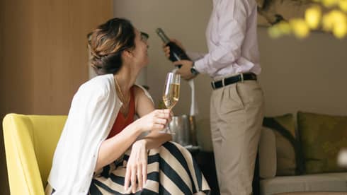 Person smiling and holding a glass of champagne while another person opens a bottle
