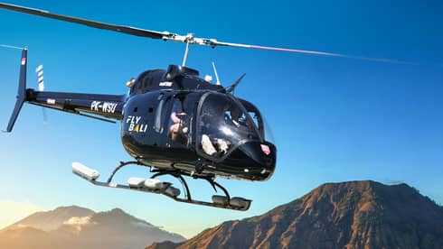 A helicopter glides across a clear blue sky past tree-lined mountains partially covered by low-lying clouds.