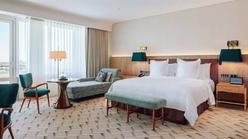 Modern hotel room with a large bed, chaise lounge, dining table, green chairs and floor-to-ceiling windows.