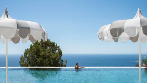 A person in an infinity-edge pool, with umbrellas in the foreground and a tree top and ocean views in the background