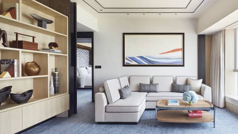 Modern hotel living area with beige sectional sofa, abstract wall art, open shelving with decor and coffee table with books and blue flowers.