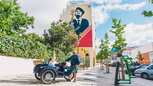 One person sits in a motorcar with another person standing next ot them on the corner of a street with a large mural in the background