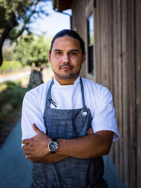 A chef in a blue apron standing outside near a wood building.