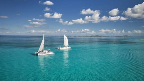 Guests aboard two catamaran sailboats glide through bright turqoise water, distant island beyond, under a cloud-dappled sky. 
