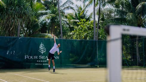 Person in athletic wear, serving on tennis court surrounded by palm trees