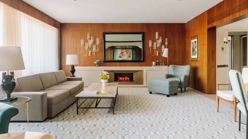 Spacious hotel living room with a sofa, armchair, fireplace, coffee table and wood-paneled walls with decorative lighting.