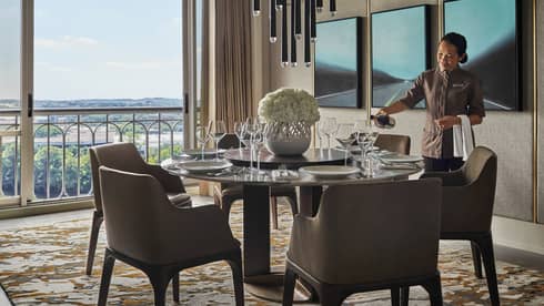 Hotel staff pours wine in glass at round dining table by floor-to-ceiling window