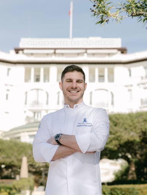 Pastry Chef Pierre-Jean Quinonero in white chef's coat, smiling with arms crossed with the Hotel in backdrop