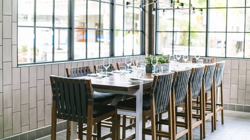 A rectangular wooden dining table with 12 black chairs under an industrial chandelier in a bright room with tiled walls. 