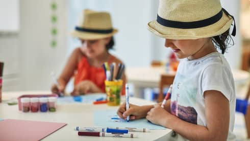 Two smiling children wearing straw fedora hats colour pictures with markers at craft table