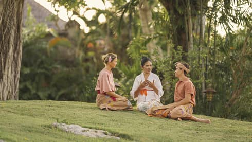 Close-up of three adults in sarongs sitting together on a grassy knoll surrounded by lush greenery and palm trees.