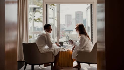 Two people in white robes sit in armchairs by an open balcony door with a cityscape view in the background, sharing a relaxed, casual conversation. 