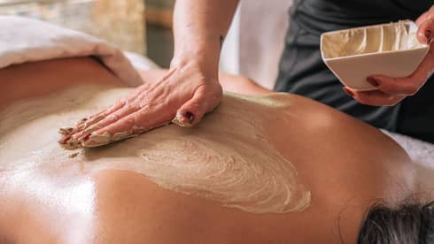 A hand rubs a sand-coloured body treatment on a person's bare back
