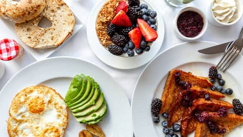 Overhead view of French toast, fried eggs, sliced avocado, granola and fruit, a bagel and various jams spread on a white table