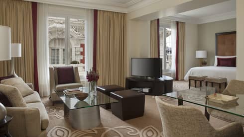 Mansion-View Junior Suite living room with beige loveseat, armchair, glass coffee table and dining table, TV, bed