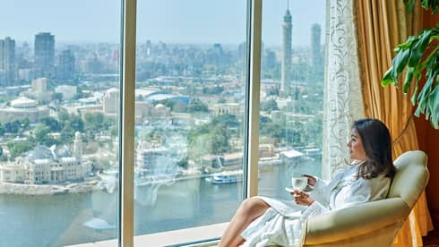 Woman wearing white bathrobe sipping coffee looks out large window over Nile River