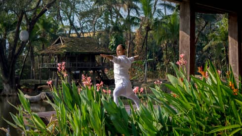 Resident yogi Dheera wearing white in yoga pose with arms outstretched by tall flowers, leaves