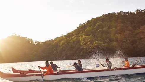 Seven people ride in a red-white-and-blue outrigger canoe with the sun setting in the distance