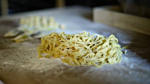 Pile of fresh tagliatelle rests on an aged wood counter covered with flour, beside scraps of pasta dough. 