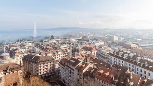A cityscape with rooftops, buildings and streets, with a large fountain shooting water into the air over a lake in the background under a clear blue sky