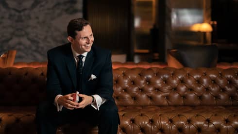 A man in a black suit sitting on a leather sofa holding playing cards.