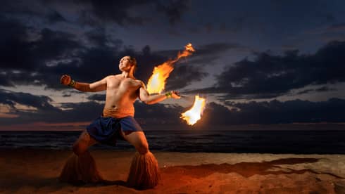 Man wearing traditional Polynesian outfit dances with torch on beach at night