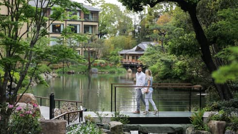 A couple leisurely walks across a footbridge, engaged in a conversation, overlooking a tranquil pond surrounded by lush greenery in a park.