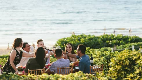 Group of friends raising glasses for a toast at an outdoor dining table by the beach, enjoying a cheerful gathering with ocean views in the background.
