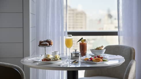 Round table with breakfast spread and two arm chairs, floor-to-ceiling windows with sheer white curtains and views of downtown New Orleans