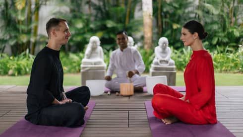 Two people sitting cross-legged on yoga mats, facing each other during a meditation session, with a spiritual guide in white and serene garden surroundings in the background.