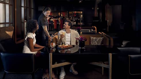 Two people wearing light-coloured cocktail attire sit on benches around a black table as a waitress brings them beverages