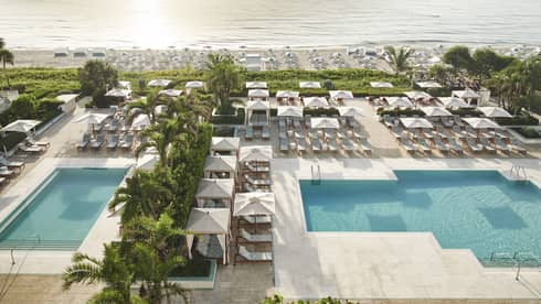 A pool area near a beach with many lounge chairs and umbrellas, surrounded by palm trees.