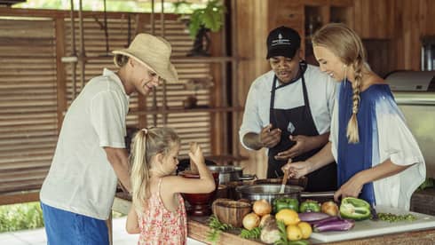 A chef instructs a guest stirring a pot, while a child uses a mortar and pestle across the counter as a third adult watches. 