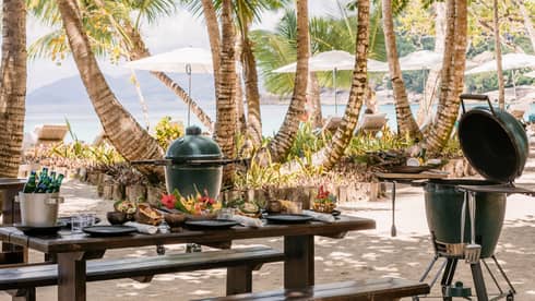 BBQ and grill equipment outside near a table and palm trees.
