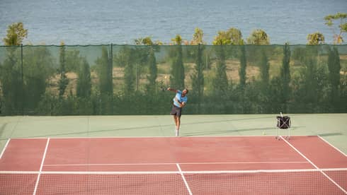 Man in blue shirt and black shorts serving a tennis ball on a seafront tennis court