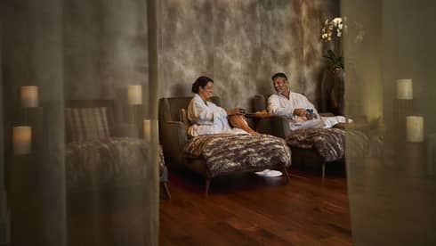 Two people wearing white spa robes relax on comfortable chairs in a serene spa space that also features a dark wood floor, candles and neutral decor.