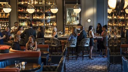 Guests and staff in a spacious, softly lit lounge space with both banquette and bar seating, hanging globe lights, and walls of shelves with bottles.