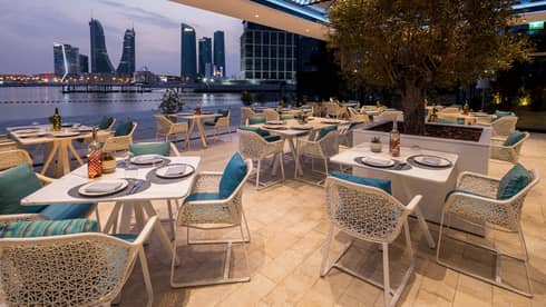 Restaurant terrace with white tables and chairs with blue cushions opens up to view of the water and a city skyline
