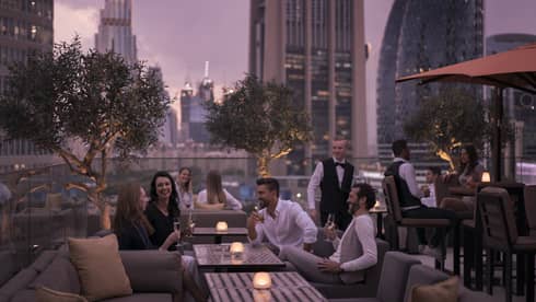 Two couples sit outdoor at Luna Bar and chat over drinks while the sun sets beyond the skulines and the sky reflects a purple hue 