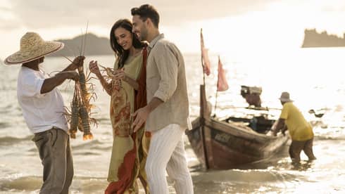 Local fisherman holds up two freshly caught lobsters for couple, traditional fishing boat in background at sunset