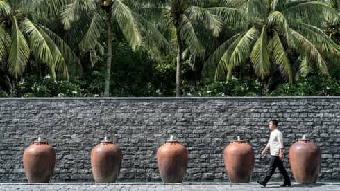 Man walks by palm trees, tall stone fence lined with large clay pot fountains