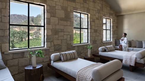 Woman in spa robe sits on couch with a tea in hand in large sunny treatment room at Spa Talisa, looking out window at Napa Valley hillside