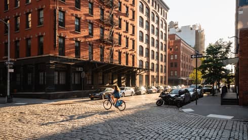 A person riding a bicycle in front of of a brick building with cars lining the street.
